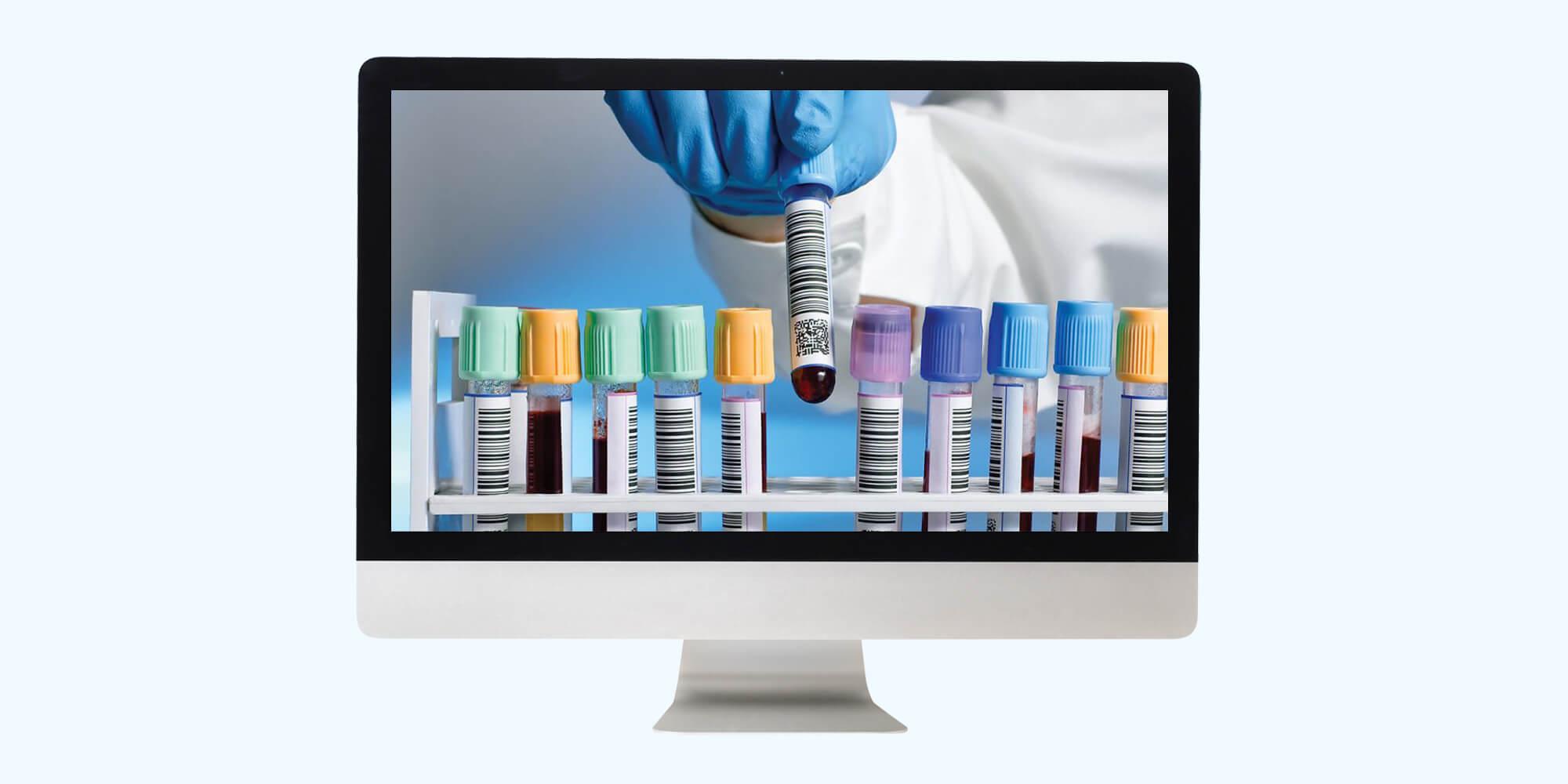 Gloved hand lifting a barcoded blood sample tube from a rack of colour-capped tubes, shown on a desktop monitor