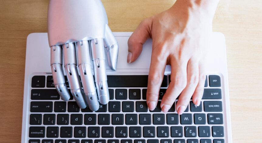 A silver robot hand and a human hand on the same laptop keyboard, seen from above