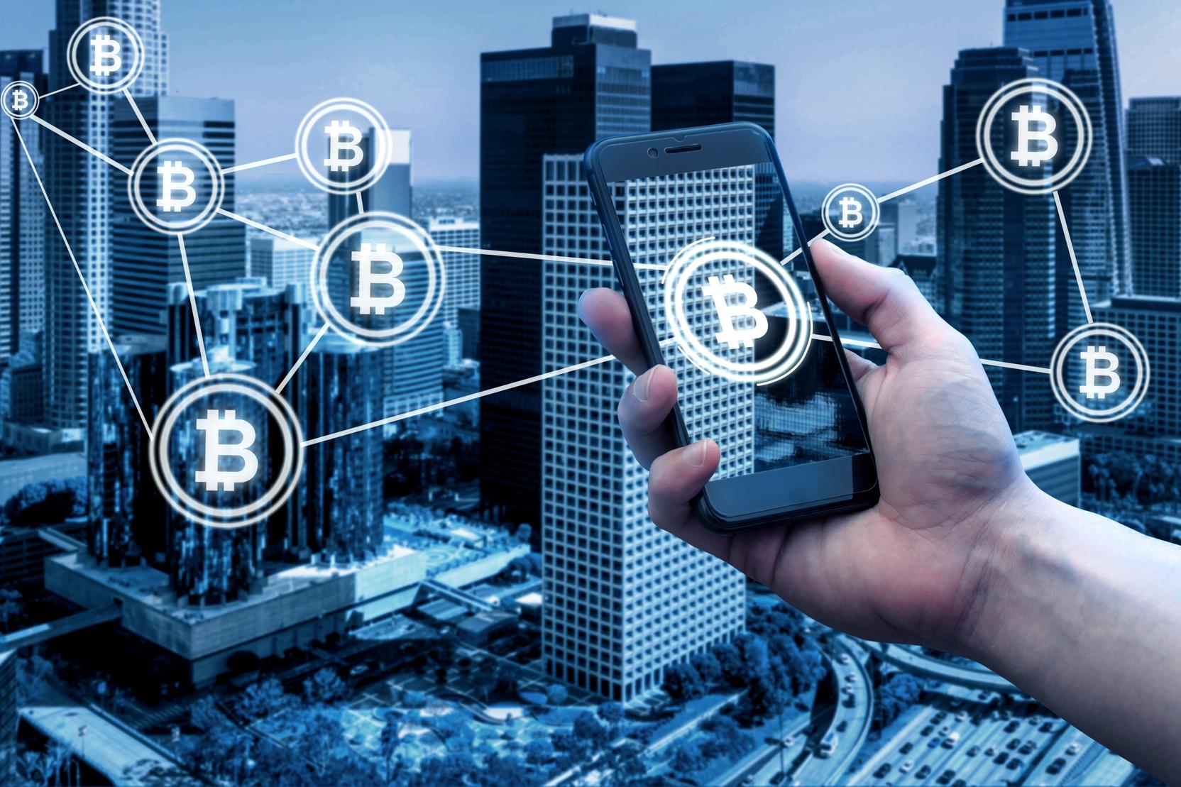 A hand holding a smartphone in front of a city skyline, with Bitcoin symbols linked by lines across the buildings